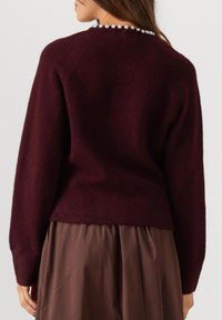 Vrouw in een kastanjebruine lange mouwen trui en een bruine rok, gezien van achteren met een parelketting om haar nek.