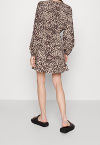 Nainen seisoo selkä kameraan päin, päällään pitkähihaiset, leopardikuosiset mini-mekot ja mustat korokepohjaiset sandaalit yksinkertaisella taustalla.