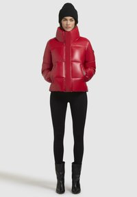 Rode pufferjack met een hoge kraag, voorzien van bovenstaande gestikte delen, ritszakken en een glanzende afwerking. Gepaard met zwarte leggings en laarzen.
