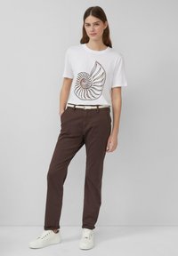 T-shirt en coton blanc avec un graphique de coquillage en spirale en marron et or. Pantalon en coton marron avec une ceinture tissée blanche. Baskets blanches.