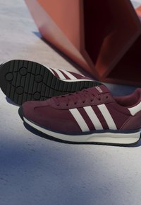 Baskets Adidas marron avec des rayures blanches et des semelles noires posées sur une surface grise à côté d'un objet géométrique rouge.