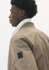Jachetă bomber maro deschis cu o textură netedă, având un guler alb și un patch logo negru pe mânecă, vedere din profil lateral.