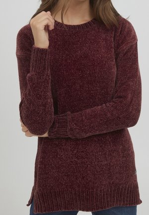 Femme portant un pull chenille bordeaux côtelé à manches longues avec un col rond, bras croisés, sur fond clair.