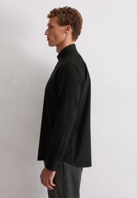 Jonge man die naar links kijkt, gekleed in een zwart shirt met lange mouwen en een hoge kraag, met grijze gestructureerde broek, voor een lichte muur.