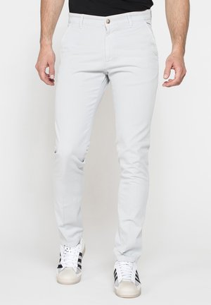 Uomo che indossa pantaloni slim fit grigio chiaro e scarpe da ginnastica bianche con strisce nere, in piedi davanti a uno sfondo semplice.