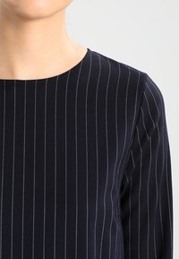 Navyblaue Bluse aus Stoff mit dünnen weißen vertikalen Streifen, rundem Ausschnitt und langen Ärmeln. Glatte Textur und taillierte Passform.