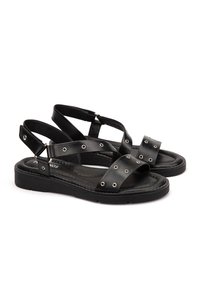 Sandales en cuir noir avec des brides croisées, accents métalliques ronds, semelle rembourrée et sangle arrière pour un maintien sécurisé. Design plat et décontracté.