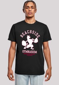 Melna kokvilnas t-kreklā ir attēlota balta un rozā grafika ar lauvu, kas ceļ svarus, ar tekstu "BEACHSIDE GYMNASIUM" virs tā.