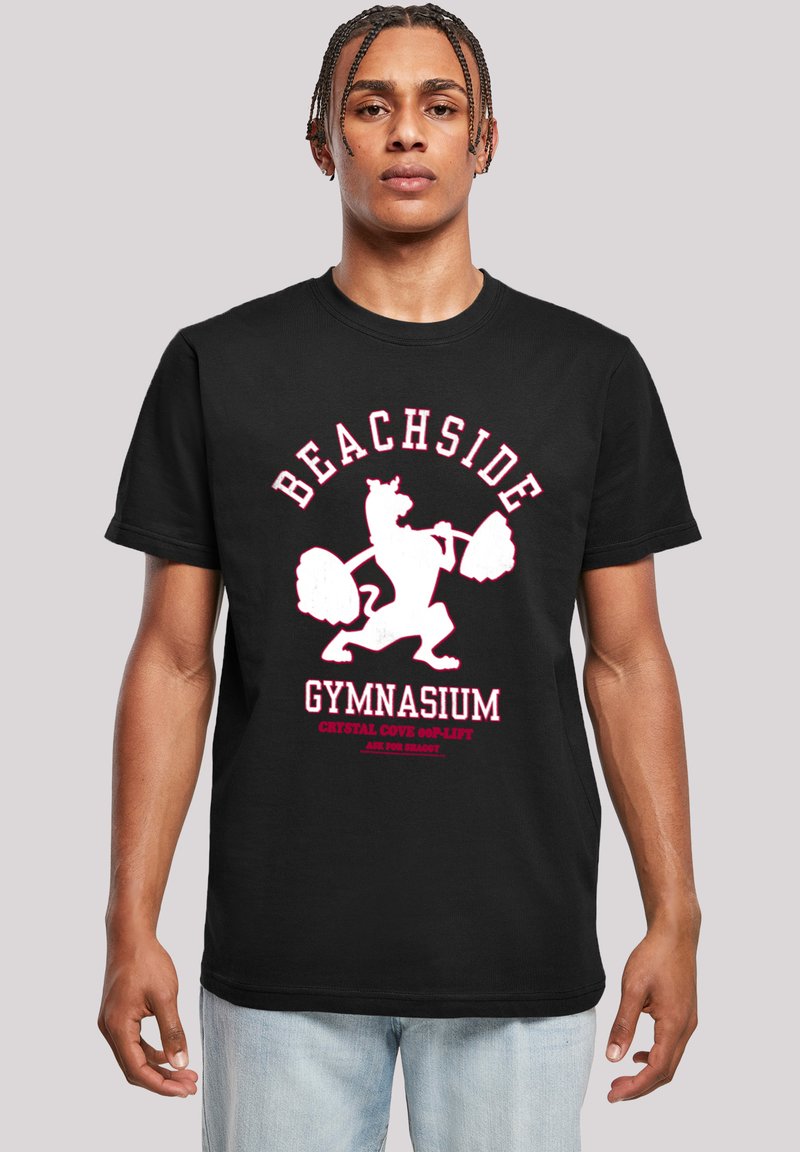 Melna kokvilnas t-kreklā ir attēlota balta un rozā grafika ar lauvu, kas ceļ svarus, ar tekstu "BEACHSIDE GYMNASIUM" virs tā.