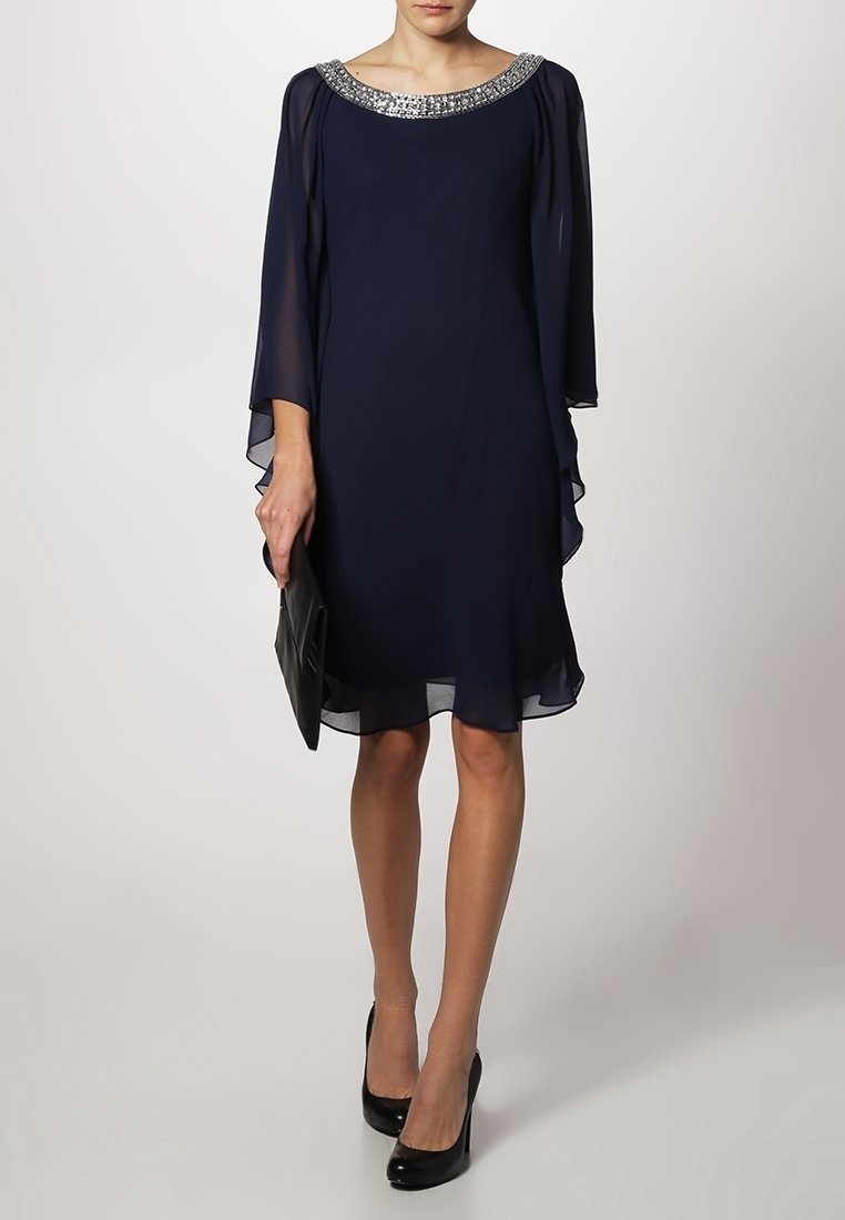 Femme portant une robe bleu marine longueur genou avec des manches transparentes et un col orné de perles, tenant une pochette noire et portant des talons noirs.