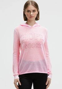 Junge Frau mit kurzem braunem Haar trägt einen hellrosa Mesh-Hoodie mit dem Text "HOPE SOCCX" und schwarze Hosen vor einem schlichten weißen Hintergrund.