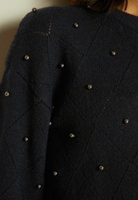 Granatowy sweter dzianinowy z wzorem w romby, ozdobiony małymi srebrnymi koralikami rozmieszczonymi na tkaninie dla dodatkowej tekstury i detali.
