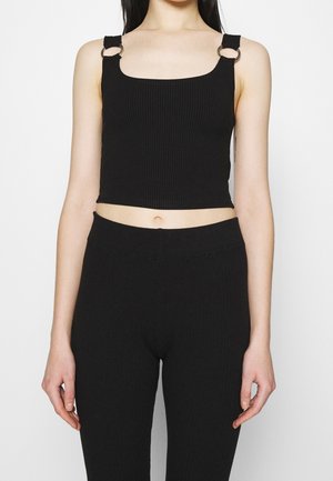 Femme portant un crop top noir à côtes avec des détails en métal sur les bretelles et un pantalon noir taille haute assorti, le tout sur fond blanc.