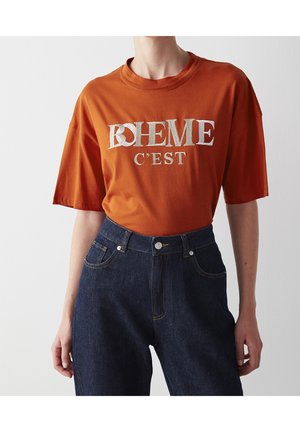 Persona con una camiseta oversize color naranja quemado con el texto "BOHEME C'EST" y jeans de tiro alto azul oscuro sobre un fondo liso.