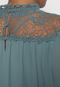Blouse turquoise avec des détails en dentelle complexe au dos, fermeture à boutons et un design plissé fluide pour plus de texture.