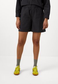 Zwarte textuurshorts met een elastische tailleband, gecombineerd met grijze sokken en gele sportschoenen. De stof heeft een subtiel reliëfpatroon.