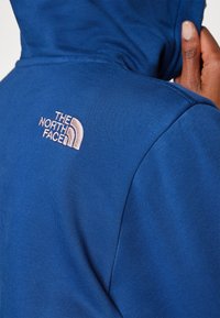 Mėlyna džemperio su gobtuvu striukė, pasižyminti minkštu audiniu, reguliuojamu gobtuvu ir rožiniu išsiuvinėtu "The North Face" logotipu ant nugaros.
