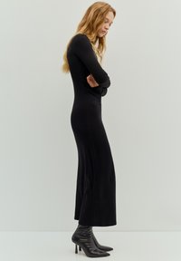 Długa, dopasowana czarna sukienka z długimi rękawami i prostą sylwetką, zestawiona z błyszczącymi czarnymi, szpiczastymi botkami na wysokim obcasie.