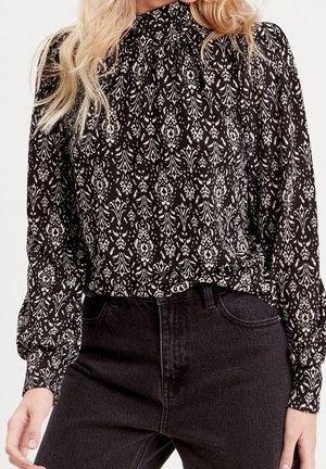 Femme portant un chemisier à motif noir et blanc à col montant avec des manches longues, rentré dans un jean noir taille haute.