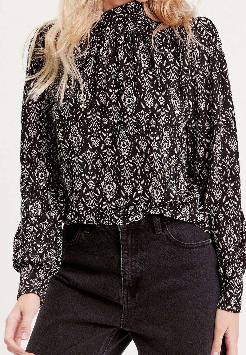 Femme portant un chemisier à motif noir et blanc à col montant avec des manches longues, rentré dans un jean noir taille haute.
