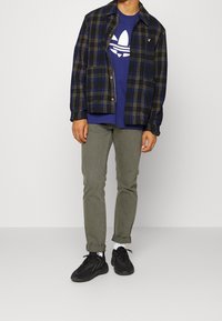 Námořnická kostkovaná bunda se žlutými akcenty přes modrou košili s bílým logem, doplněná olivově zelenými kalhotami a černými teniskami.