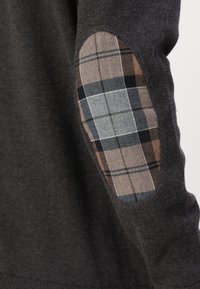 Pulover gri închis, cu țesătură texturată și petice pe coate în carouri, în nuanțe de maro, gri și negru, evidențiind un element de design contrastant.
