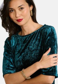 Žena s tmavými vlasmi, ktorá má na sebe červenú rúž, zlaté textúrované náušnice, zodpovedaj�úci náramok a tyrkysový zamatový top s trojštvrťovými rukávmi.