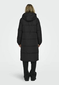 Manteau long noir en duvet avec capuche, présentant un design matelassé, un large col et des poignets élastiques. Le matériau semble résistant à l'eau et durable.