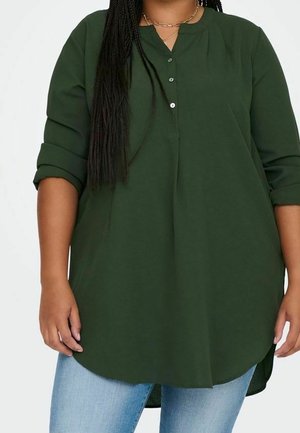 Tunique manches longues vert foncé avec manches retroussées, col boutonné, portée avec un jean bleu clair et des colliers dorés superposés.