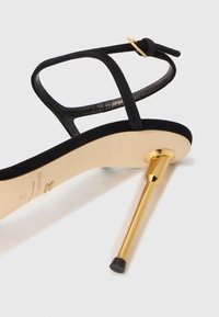 Black suede sandal with thin straps, a gold-tone heel, and a beige sole. Features a smooth texture and a pointed heel design.