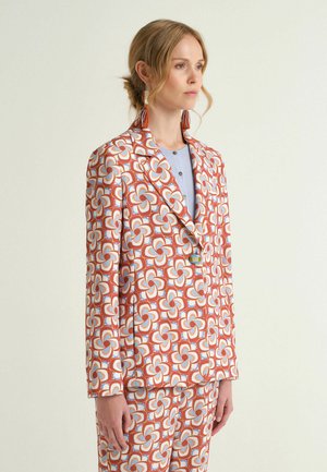 Frau in einem farbenfrohen Anzug mit orangefarbenen, blauen und weißen Blumenmustern, die orangefarbene Quastenohrringe trägt und vor einem schlichten Hintergrund steht.