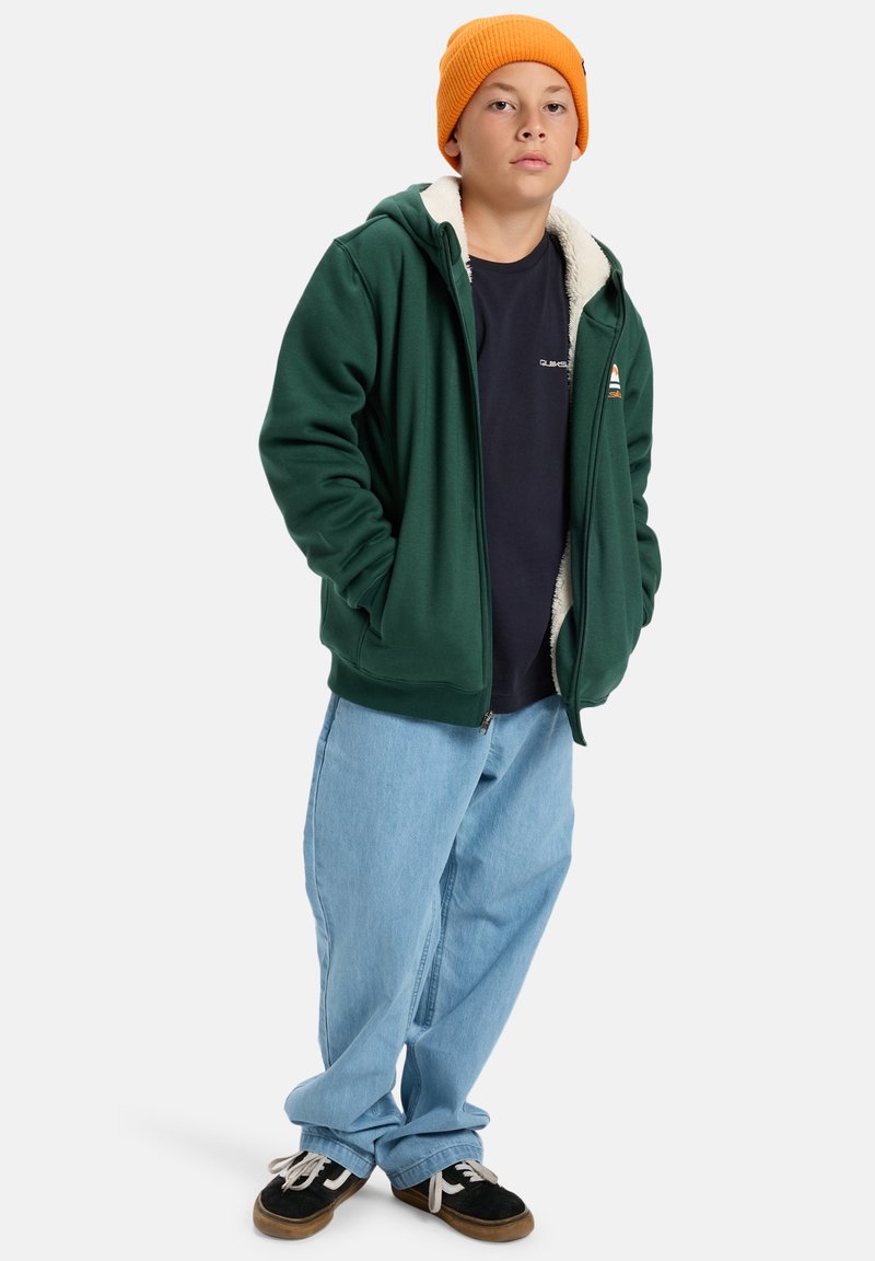 Sudadera verde con cremallera y forro de forro polar, usada sobre una camiseta azul marino, combinada con jeans holgados de color azul claro y zapatos negros. Gorro naranja en la cabeza.