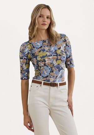 Langermet bluse med blomsterdesign i blått, gult og brunt; tettsittende design; glatt stoff; kombinert med hvite jeans og et brunt belte.