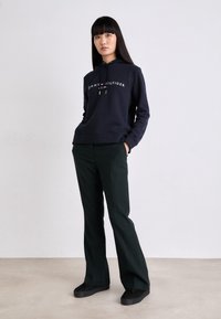 Sudadera navy con logo blanco, corte holgado y cordón ajustable. Pantalones verdes oscuros, de corte recto. Zapatos negros con suelas gruesas. Fondo de estudio sencillo.