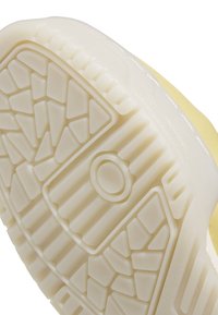 Suela de zapatilla amarilla con goma blanca texturizada que presenta patrones triangulares y rectangulares, detalle de logotipo circular y acentos en el borde elevado.