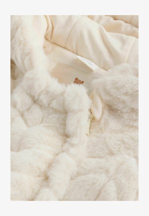 Interiér krémového umělého kožíšku s měkkou, nadýchanou texturou, který má malý detail medvídka a částečně viditelnou látkovou etiketu.