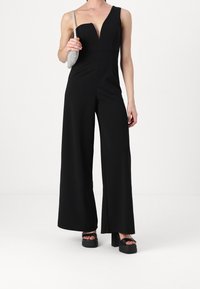 Svart ärmlös byxdress med djup V-ringning, vida ben och figurnära midja. Matchad med en metallisk axelväska och plattformssandaler.