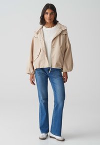 Giacca beige leggera con cappuccio, bottoni a pressione e tasche laterali. Abbinata a jeans a zampa d'elefante azzurri chiari e sneakers bianche. Maglione bianco con texture sotto.