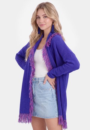 Jonge vrouw met lang blond haar draagt een blauwe cardigan met franjes, een wit hemdje, een lichte denim rok en blauwe kwastjes oorbellen, poseert met hand in de zij.