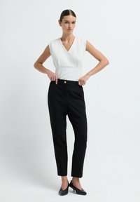 Vrouw past zwarte hoog-waist tailored broek with gouden knoop aan, draagt een mouwloze witte V-hals top en zwarte hakken tegen een effen achtergrond.