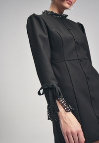 Robe noire avec un faux col haut. Caractéristiques des manches bouffantes et un détail à pois aux poignets, attachés avec un ruban noir. Texture lisse.