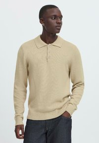 Pulover tricotat bej cu guler clasic, având trei nasturi și manșete și tiv cu nervuri. Textură netedă, design simplu, potrivire lejeră.