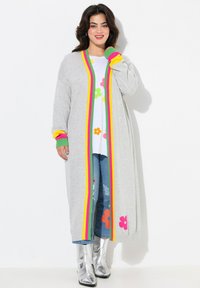 Grijze lange cardigan met kleurrijke strepen in roze, geel en groen. Draagt over een witte top met bloemenpatronen, gecombineerd met blauwe jeans en zilveren laarzen.