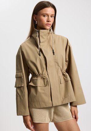 Beżowa wodoodporna kurtka z wysokim kołnierzem, talią ze sznurkiem, dwoma bocznymi kieszeniami i elastycznymi mankietami. Gładka faktura i luźny fason.