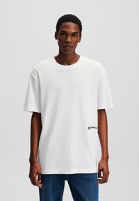 Biela bavlnená tričko s krátkymi rukávmi, vo voľnom strihu, okrúhlym výstrihom a malým čiernym logom na spodnej ľavej strane.