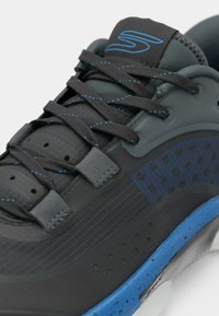 Chaussure de sport gris foncé avec une tige texturée, des accents bleus et des détails en mesh. Elle présente des lacets noirs et des sections à motifs sur les côtés.