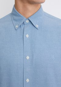 Light blue button-up shirt with a collared neck, made from a soft fabric, featuring white buttons and a subtle embroidered logo on the chest.