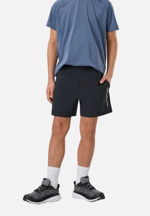 Person iført en blå kortærmet skjorte, sorte træningsshorts med sidelomme, hvide mellemlange sokker og sorte løbesko, stående afslappet.