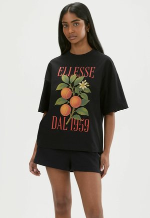 Frau trägt schwarzes oversized T-Shirt mit Orangenfrucht- und grünen Blättern Design, rotem Schriftzug "ELLESSE DAL 1959", kombiniert mit schwarzen Shorts.