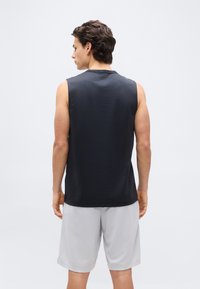 Νέος άνδρας που στέκεται με την πλάτη προς την κάμερα, φορώντας ένα μαύρο αμάνικο πουκάμισο και ανοιχτό γκρι σορτς σε απλό φόντο.